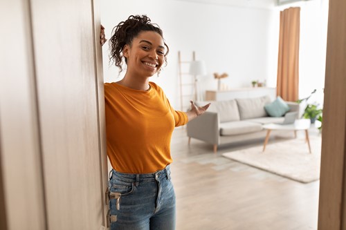 En afroamerikansk kvinna hälsar besökare vid dörröppningen och visar sin lägenhets vardagsrum.