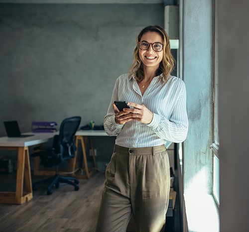 A smiling woman standing by the window in her office holding a mobile phone.